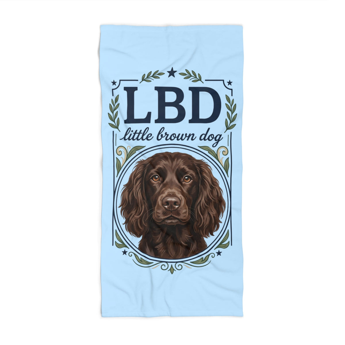 A light blue towel featuring a printed design of a brown spaniel dog inside an oval frame, with the text 'LBD little brown dog' and decorative green leaves and stars.